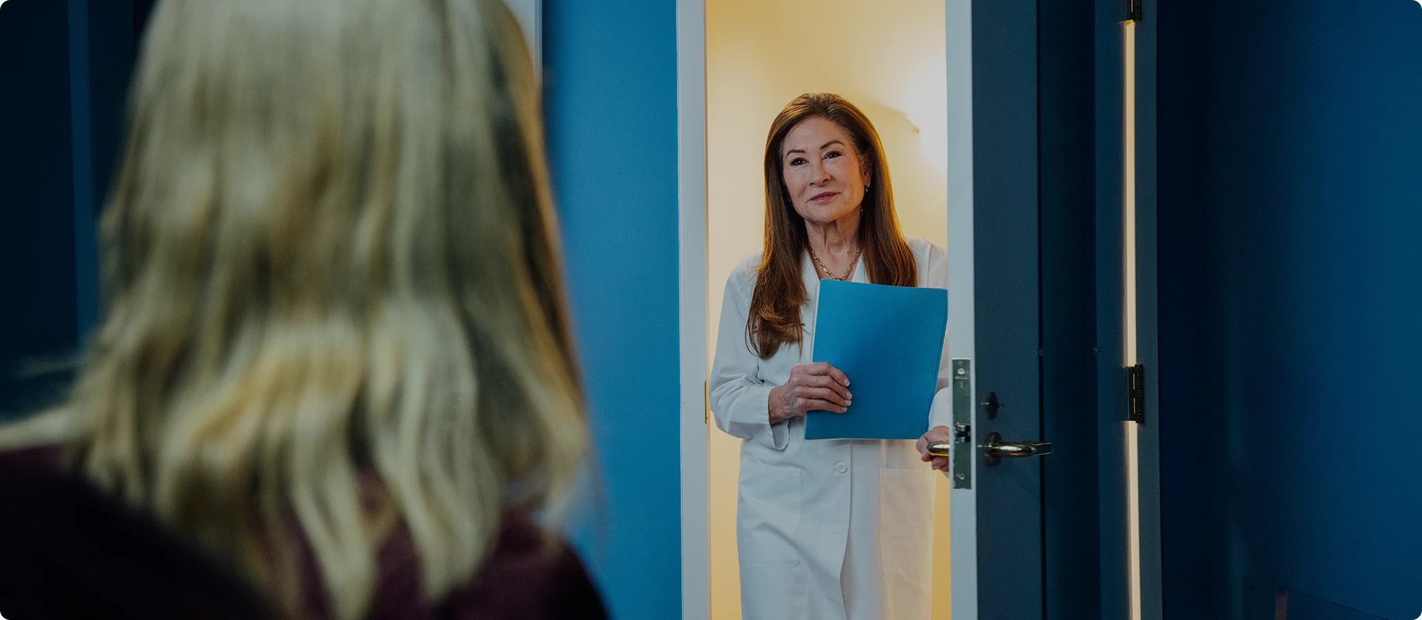 Woman with blue folder standing in doorway speaking to blonde person viewed from behind