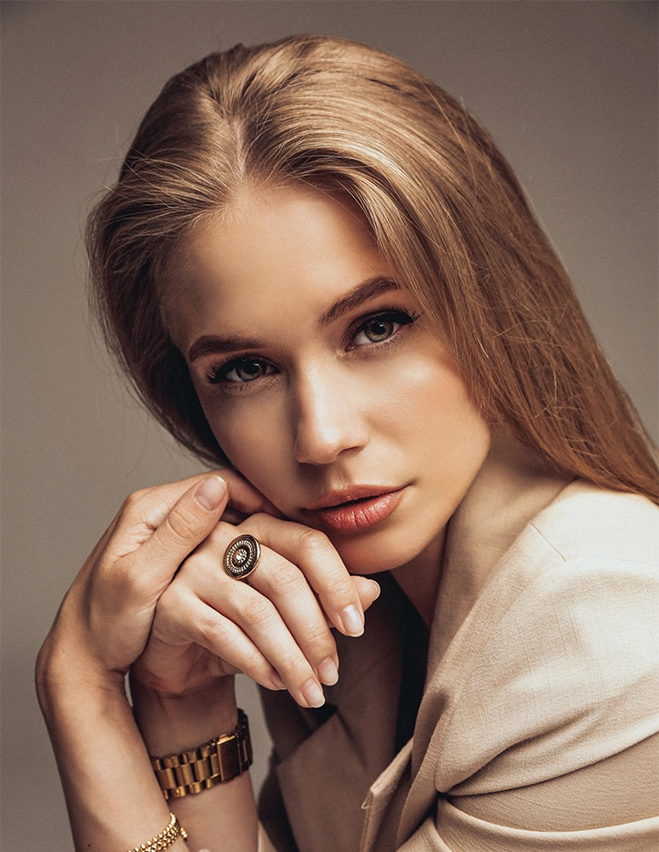 Elegant blonde woman in beige outfit wearing statement ring and gold bracelet posing with hands near face