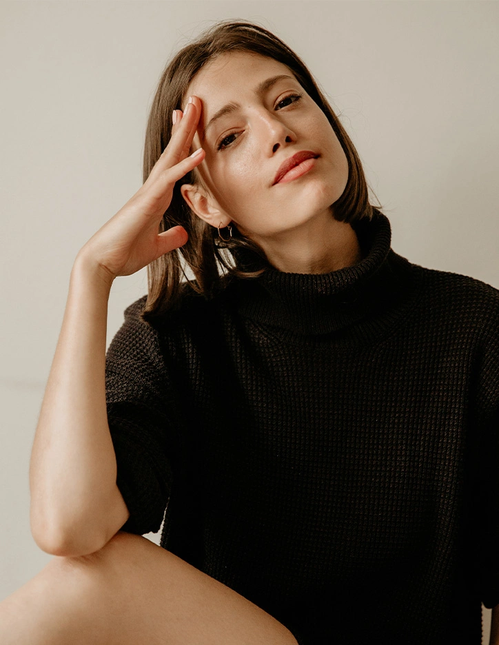 Woman in black turtleneck sweater posing with hand near face in contemplative portrait against neutral background