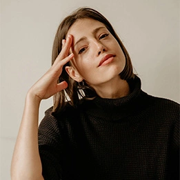Woman in black ribbed sweater posing with hand near face against neutral background