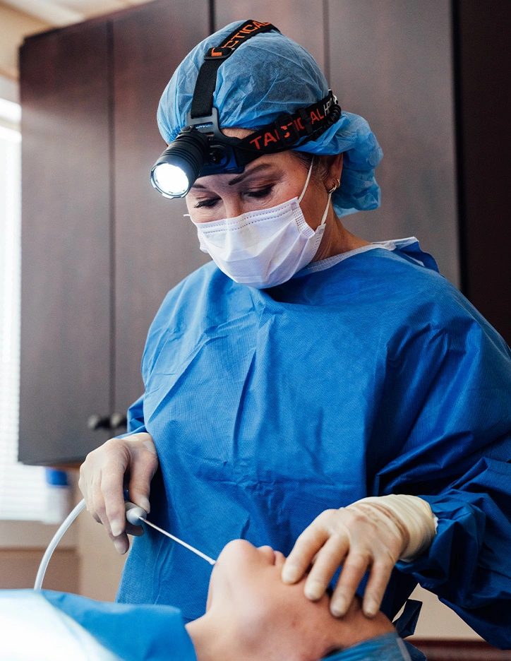 Dentist in blue surgical gown and mask performing dental procedure with headlamp and instruments