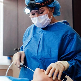 Laurie A. Casas, MD in blue scrubs and surgical mask performing medical procedure with headlamp in operating room