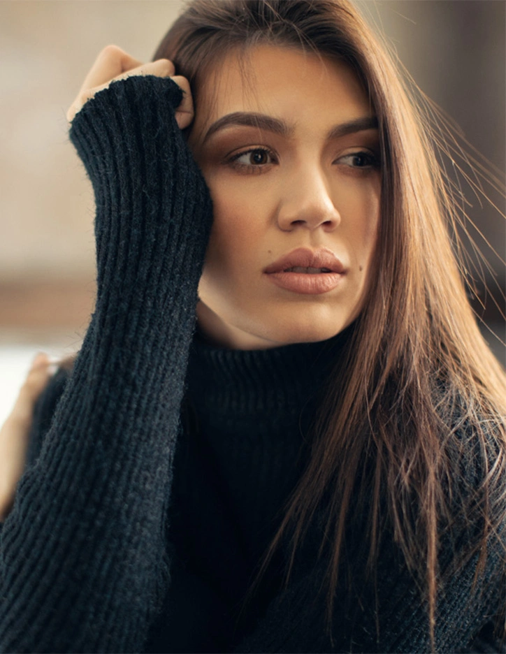Woman with long brown hair wearing black turtleneck sweater touching her head in natural light