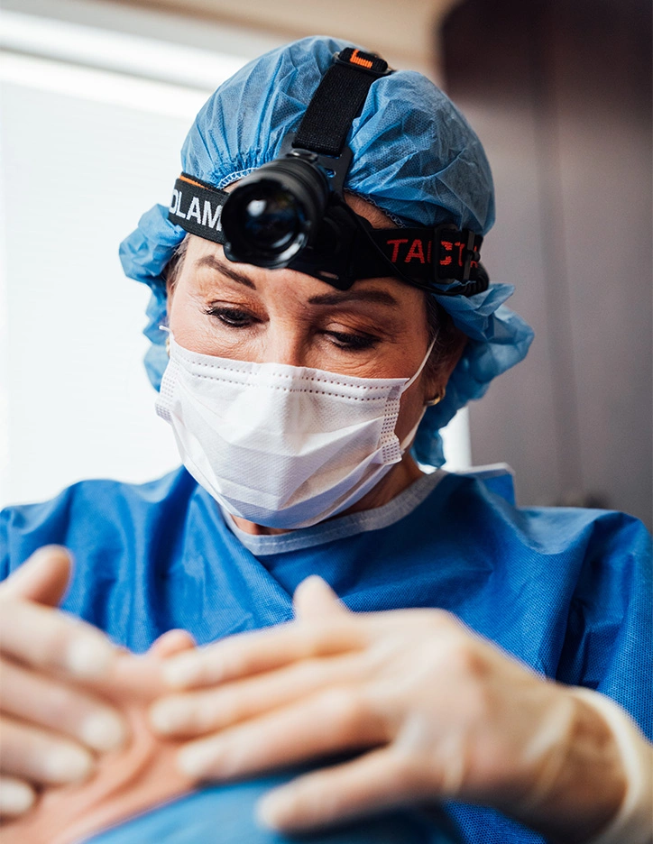 Surgeon wearing blue scrubs, surgical mask, and headlamp examining patient's hand during medical procedure
