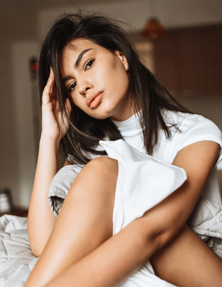 Young woman with dark hair in white shirt sitting on bed looking at camera in bedroom setting