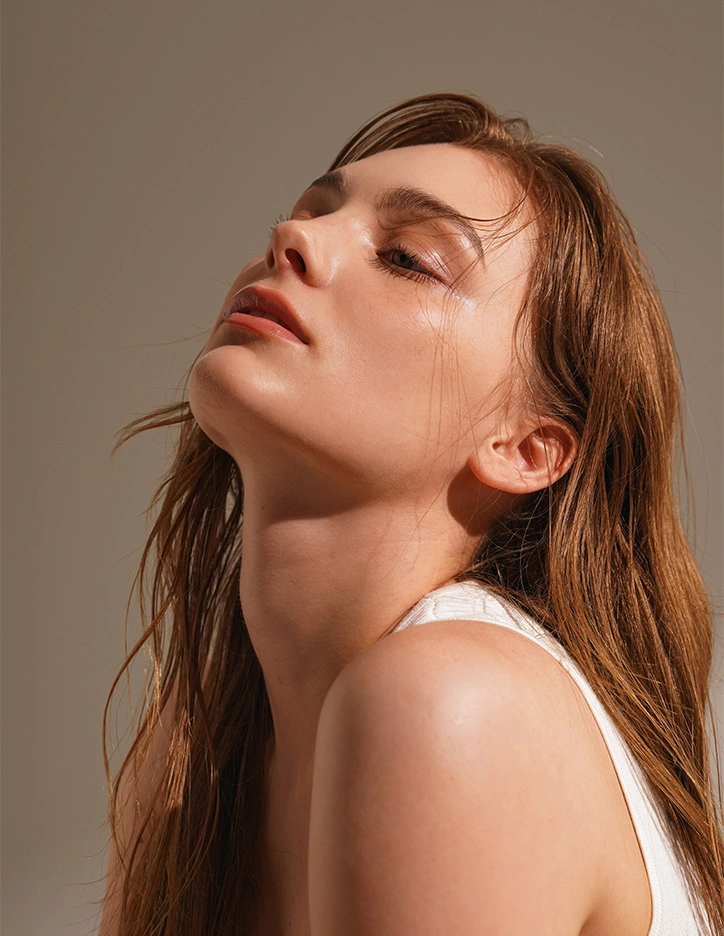 Woman with glowing skin and natural makeup tilting head back against neutral background in beauty portrait