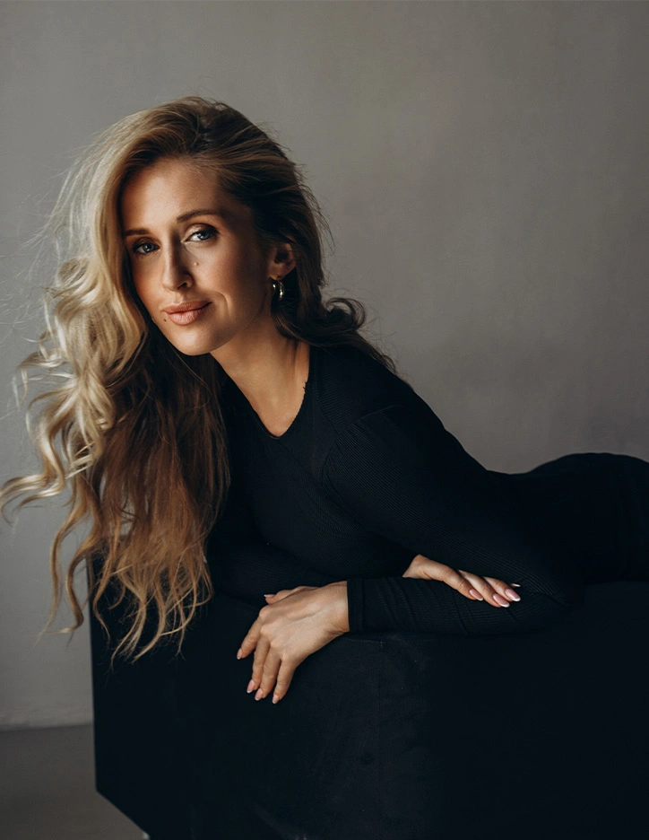 Woman with long blonde hair wearing elegant black dress posing against neutral gray background in professional portrait
