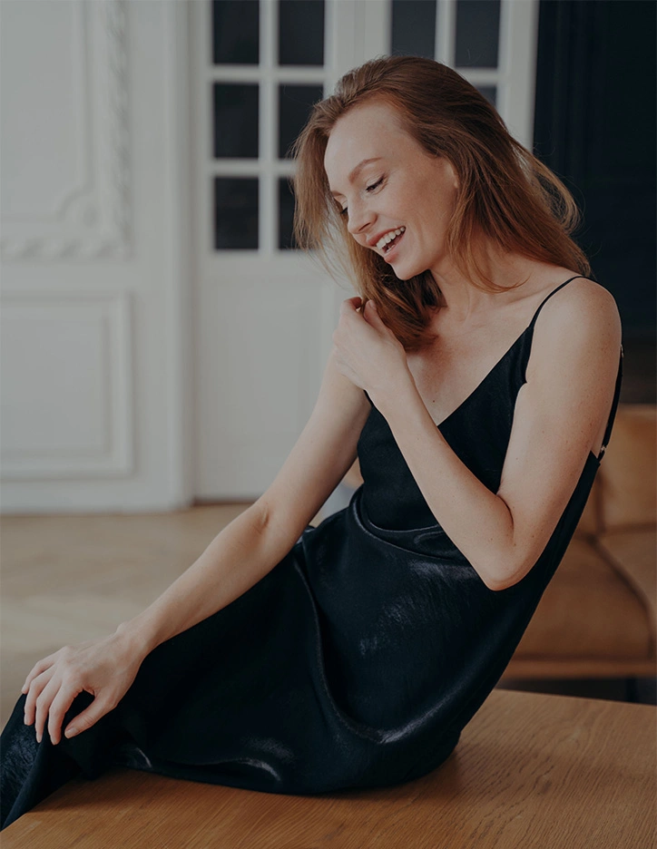 Woman in elegant black dress sitting on floor laughing joyfully in bright interior room