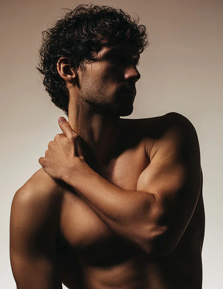 Shirtless man with curly hair embracing himself against neutral background, self-care and body confidence concept