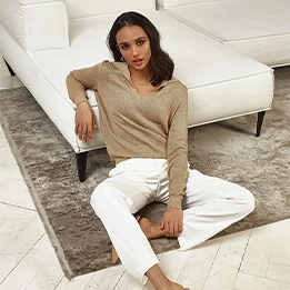 Woman wearing beige knit sweater and white jeans sitting on floor beside white couch in modern living room